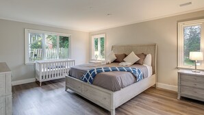 Ein Schlafzimmer mit einem Bett, einem Babybett, einem Nachttisch und einem Fenster mit Blick auf Grünflächen.