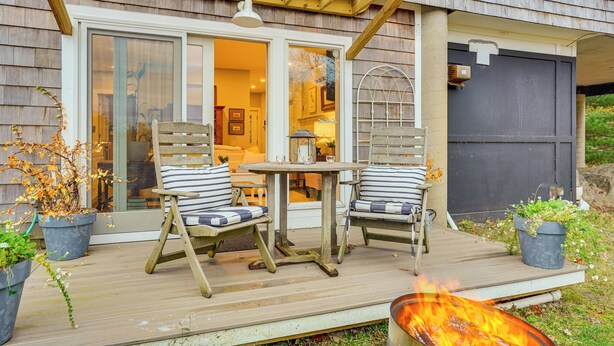 A wooden deck with two striped chairs and a small table, a fire pit, and a house with a garage door. - Airstream & Ocean View: Unique Gloucester Getaway (Gloucester)
