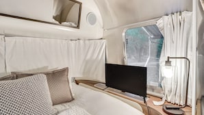A compact, modern RV interior with a bed, a mounted TV, and a window with curtains. - Airstream & Ocean View: Unique Gloucester Getaway (Gloucester)