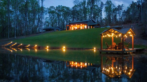 A lakeside lodge with a reflective pond, a covered seating area, and a main building with multiple lights.