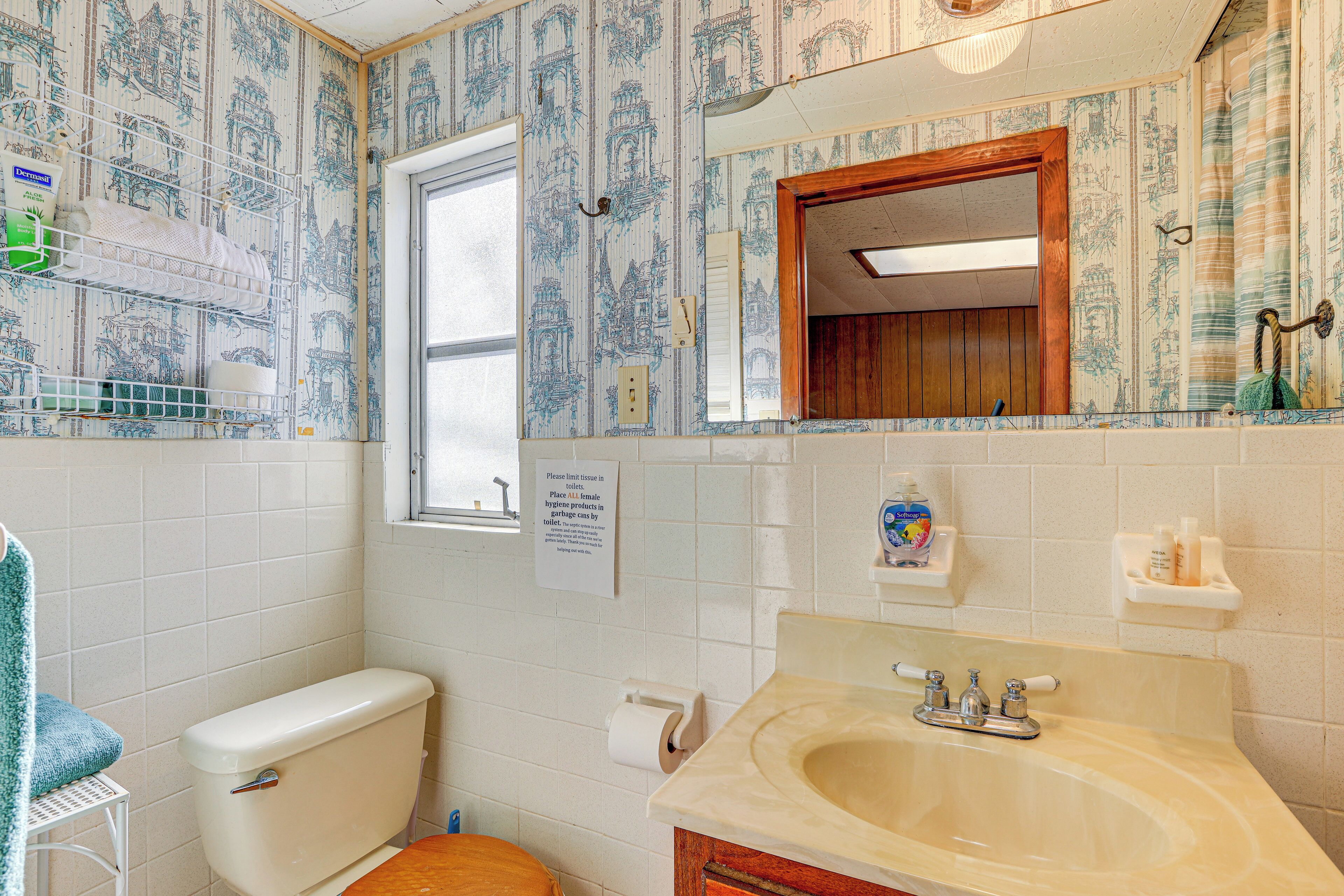 A bathroom with a toilet, sink, and mirror. There is a window and a sign on the wall.
