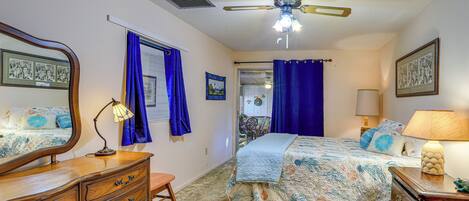 A bedroom with a bed, dresser, mirror, ceiling fan, and a view into another room.