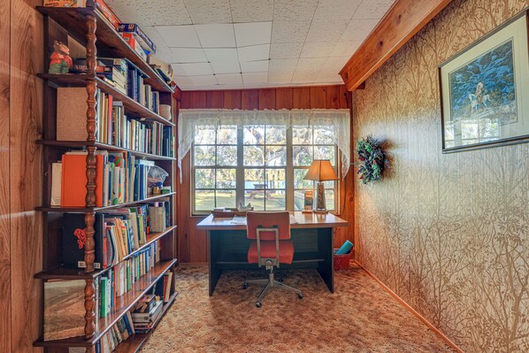 Une pièce avec un bureau en bois, une bibliothèque, une fenêtre donnant sur des arbres et un tableau encadré au mur.