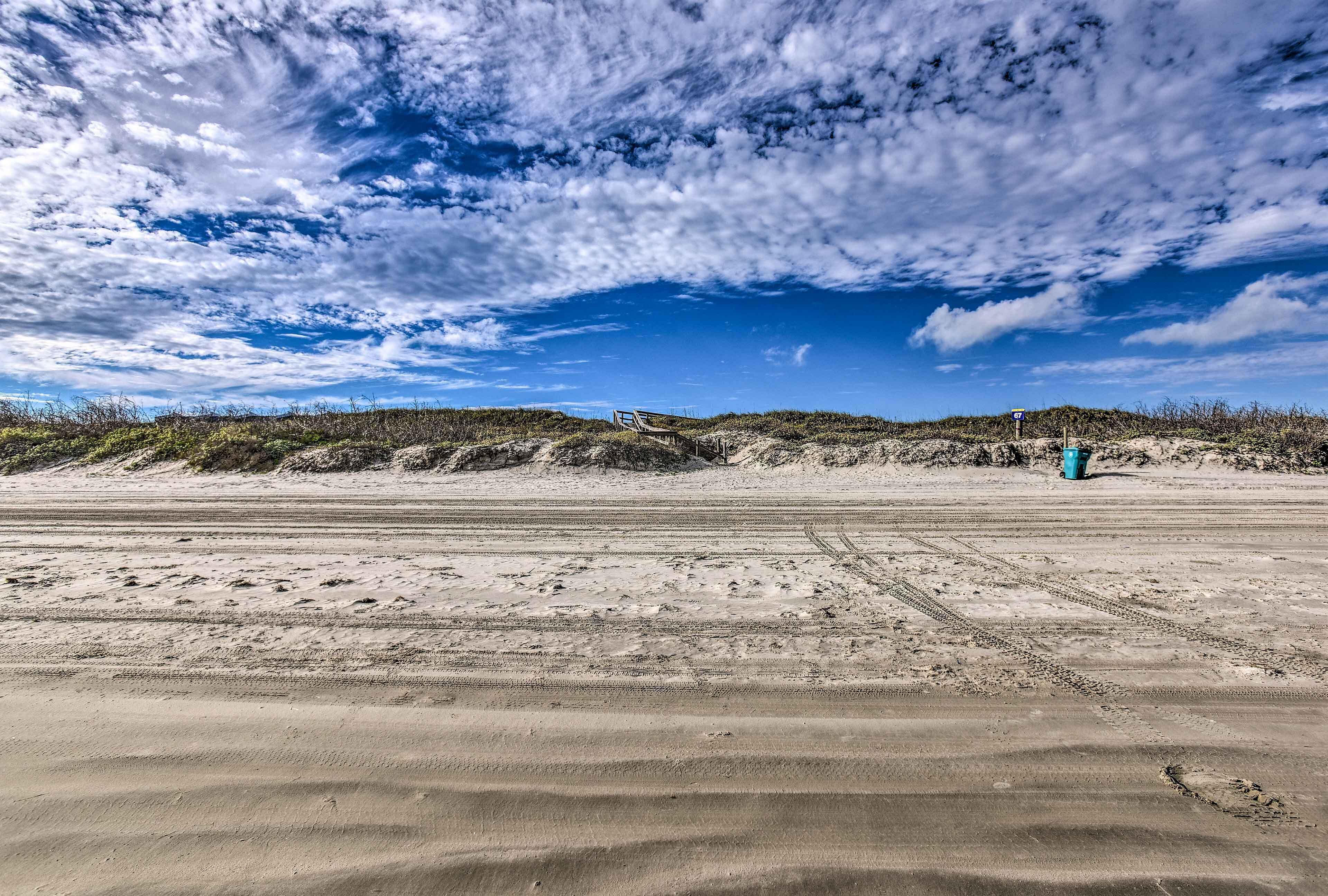A sandy beach with tire tracks and a blue trash bin.