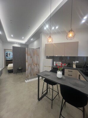 Une cuisine moderne avec un coin repas, comprenant un îlot central, un éclairage suspendu et une vue sur la chambre adjacente.