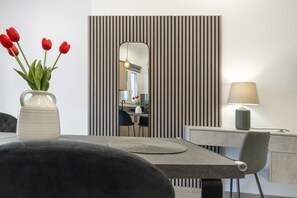 Un espace repas moderne comprenant une table, des chaises, un vase avec des fleurs rouges, une lampe et un panneau mural à rayures.
