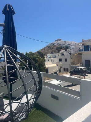 A white building with a balcony, a blue umbrella, and a view of a hill with white buildings.