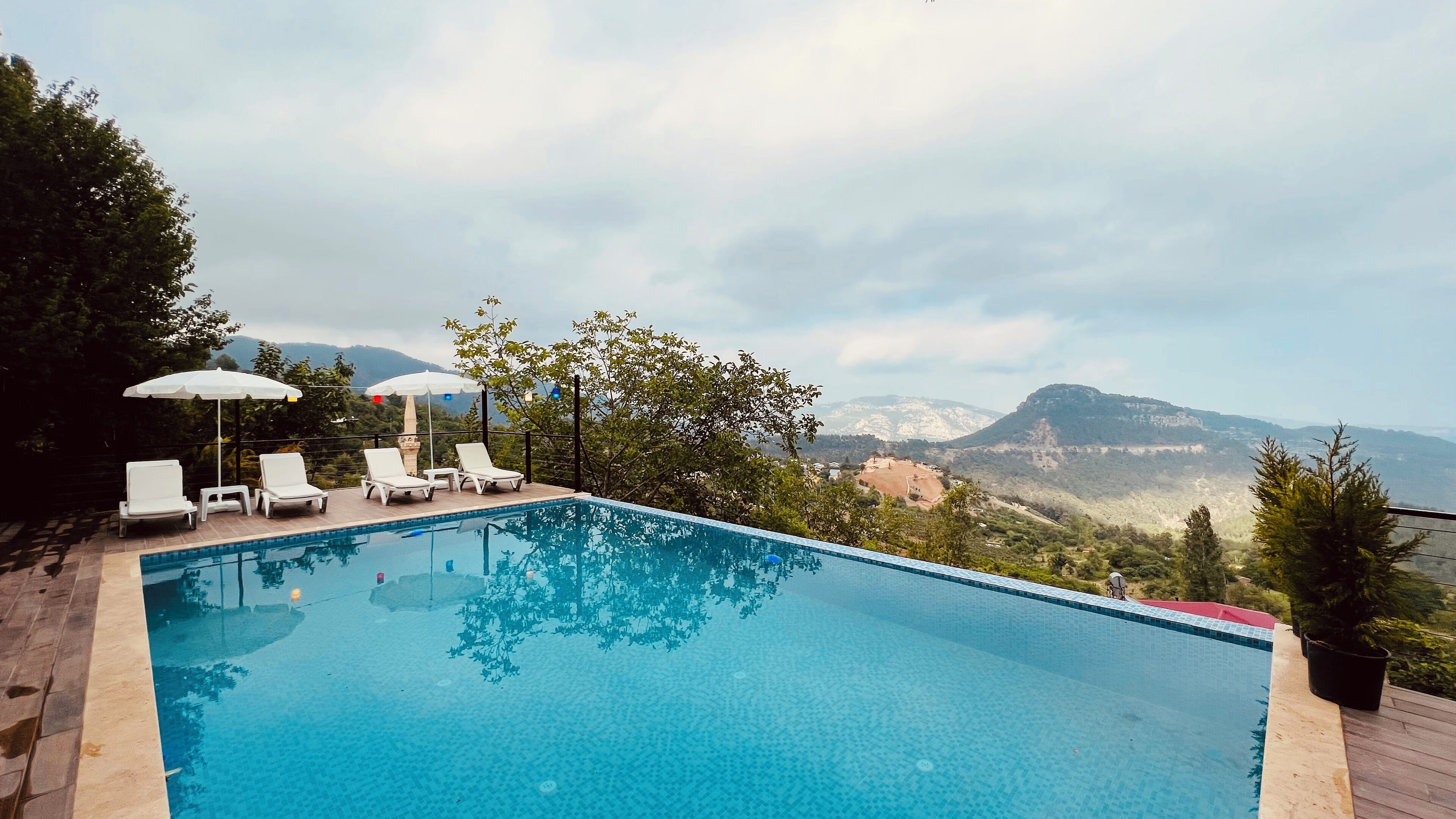 Piscina con sillas de descanso blancas y sombrillas, rodeada de vegetación y montañas al fondo.