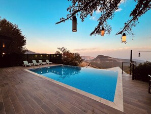 A rectangular infinity pool with a wooden deck, surrounded by lounge chairs and string lights. - Otelox Sinap Villa (Erdemli)