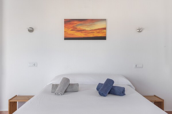 A bed with white bedding, two rolled towels, a painting of a sunset, and a wooden nightstand.
