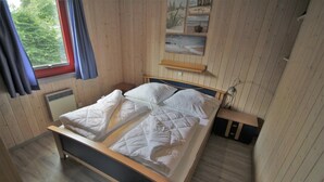 A wooden-paneled room with a bed, a window with curtains, and a wall-mounted shelf. - Ferienhaus im Ferienpark Mirow (Mirow)