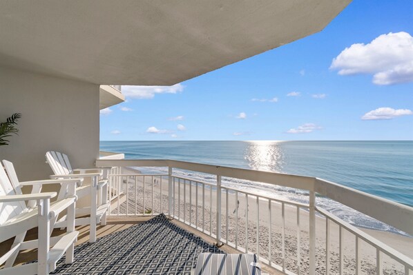 A balcony with white chairs and a patterned rug, overlooking a beach with the ocean and a clear sky.