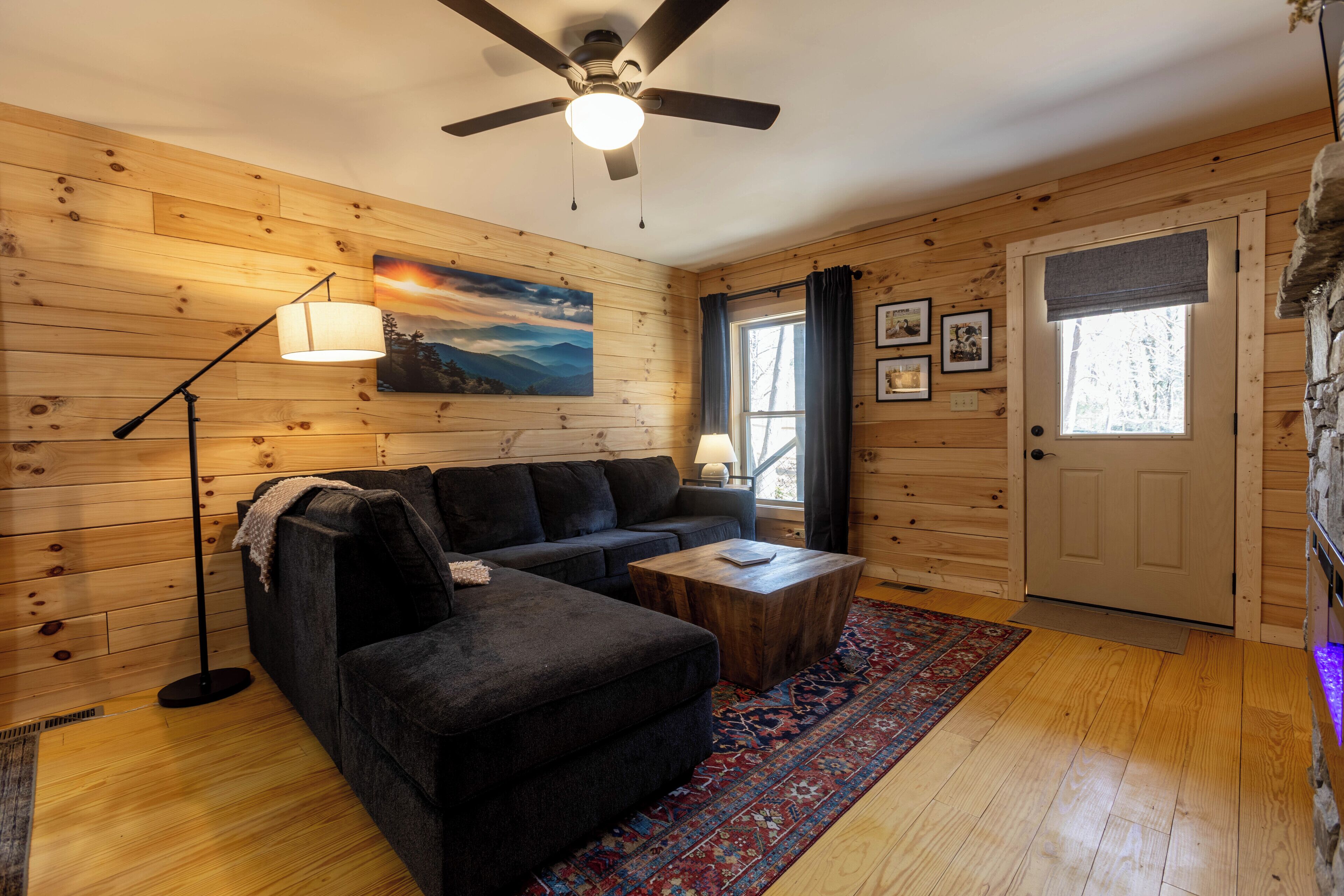 A cozy room with wooden walls, a ceiling fan, a black sofa, a wooden coffee table, a patterned rug, and a door with a window.