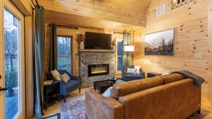 Accogliente interno di una baita con camino in pietra, pareti in legno, una grande TV e un'area salotto confortevole.