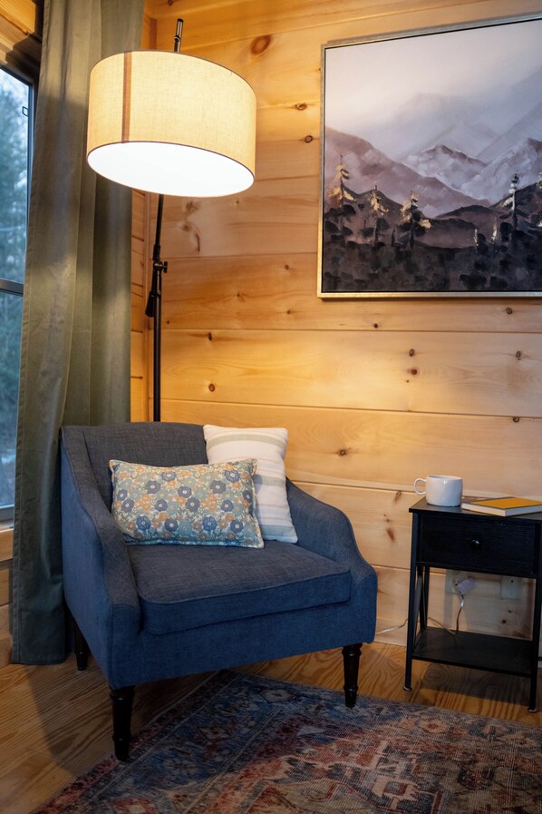 A cozy room with a blue armchair, patterned pillow, and a lamp. A painting of mountains hangs on the wall, and a small table with a cup and book is visible.