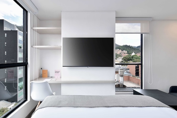 A modern hotel room with a large window, a flat-screen TV mounted on the wall, a desk, and a bed with a gray bedspread.