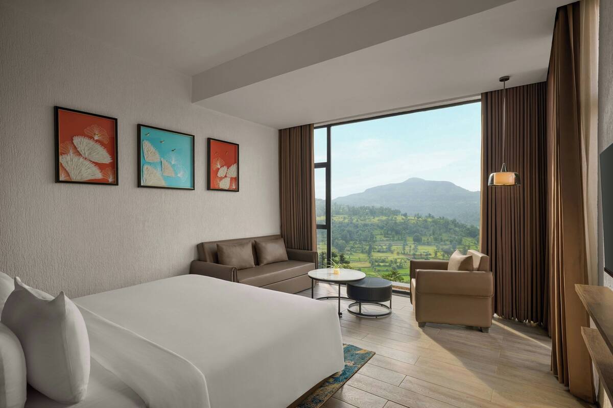 Ein Hotelzimmer mit einem großen Bett, einer Sitzecke, einem kleinen runden Tisch und Blick auf eine gebirgige Landschaft durch ein großes Fenster.