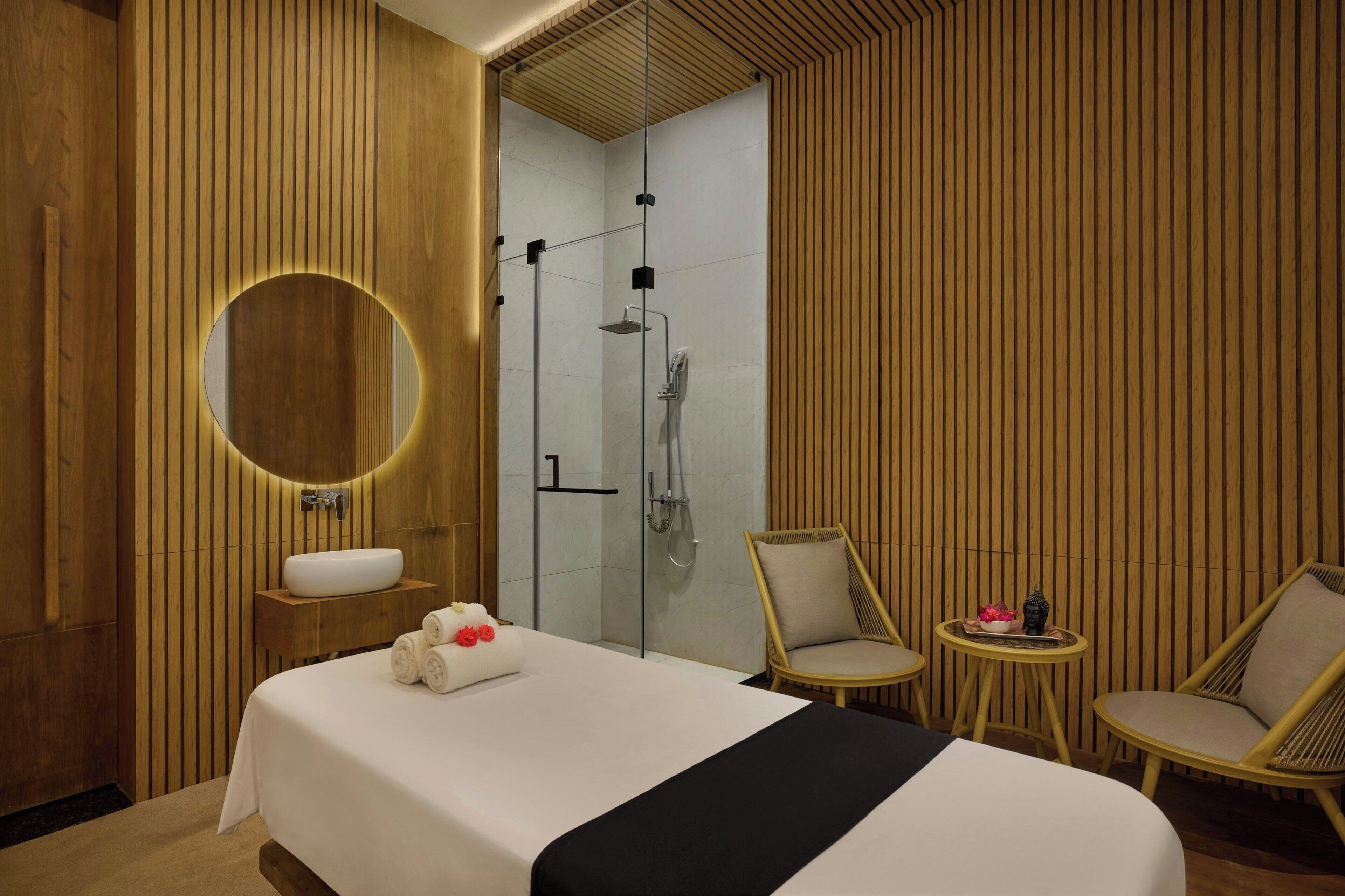 A modern hotel room with a large bed, a round mirror, a sink, and a shower. There are two wicker chairs and a small table with a vase.