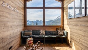 A wooden cabin interior with a large window offering a mountain view, a wooden bench, and a rocking chair.