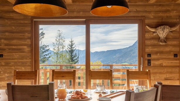 A wooden cabin with a dining table set for breakfast, a large window with a mountain view, and a wooden bull head decoration on the wall.