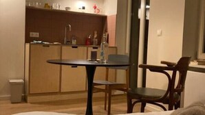 Une chambre d’hôtel avec un lit, une petite table et une chaise. Au fond de la pièce, on aperçoit une kitchenette équipée d’un évier, d’une cuisinière et de placards.