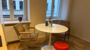 Una habitación con una mesa redonda blanca, dos sillas, un taburete rojo, un tapete tejido y una ventana con vistas a edificios.