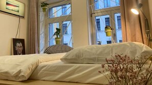 Un dormitorio con una cama, dos ventanas, una planta, una lámpara y un cuadro en la pared.