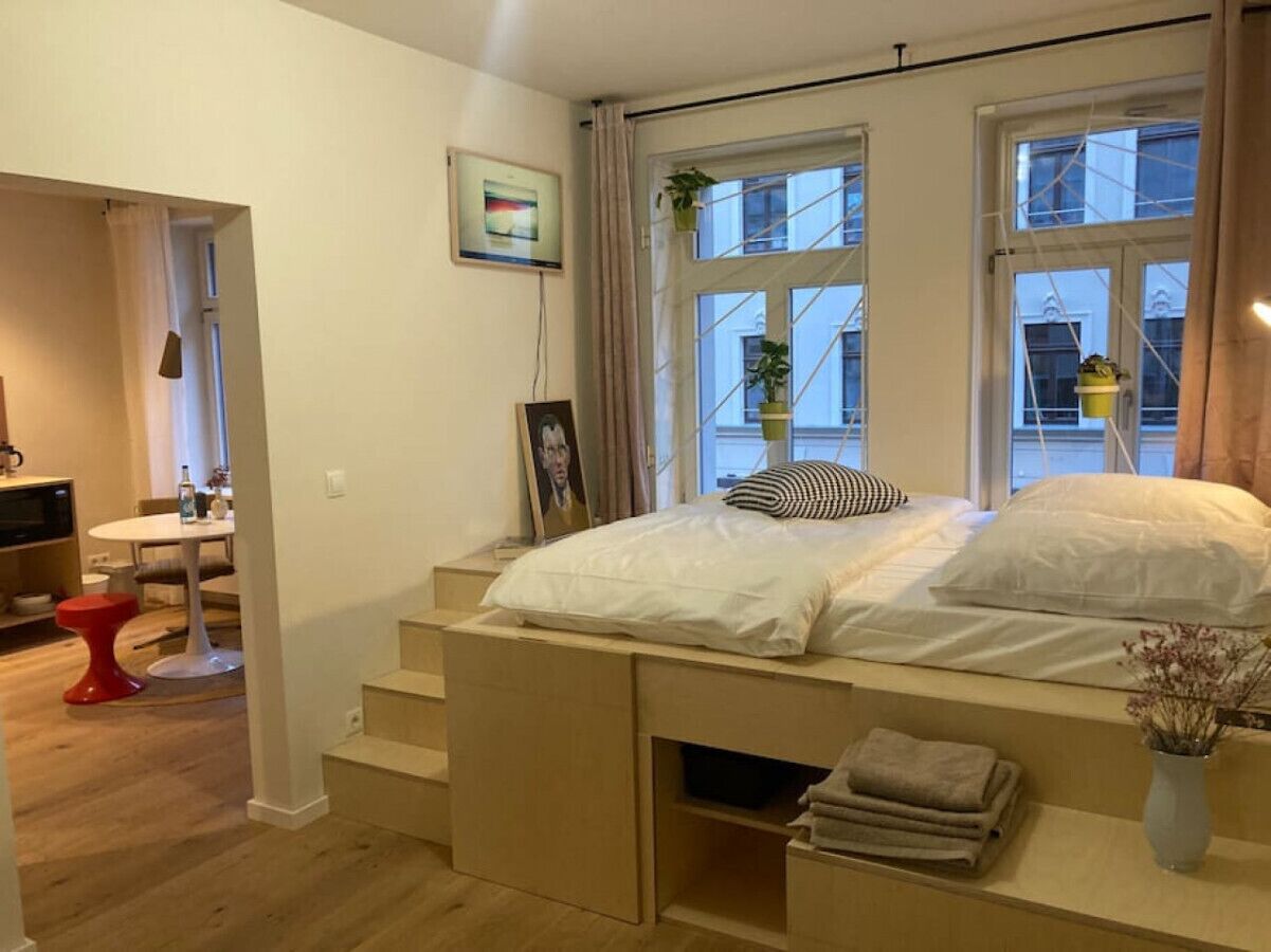 A bedroom with a bed, a desk, and a chair. There are windows with curtains and a plant on the windowsill.