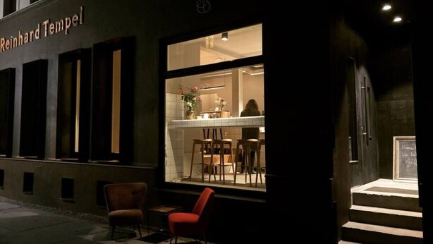 Un restaurante con fachada oscura y un interior muy iluminado. Hay dos sillas frente a la entrada, y se observa un menú en una pizarra en el interior.