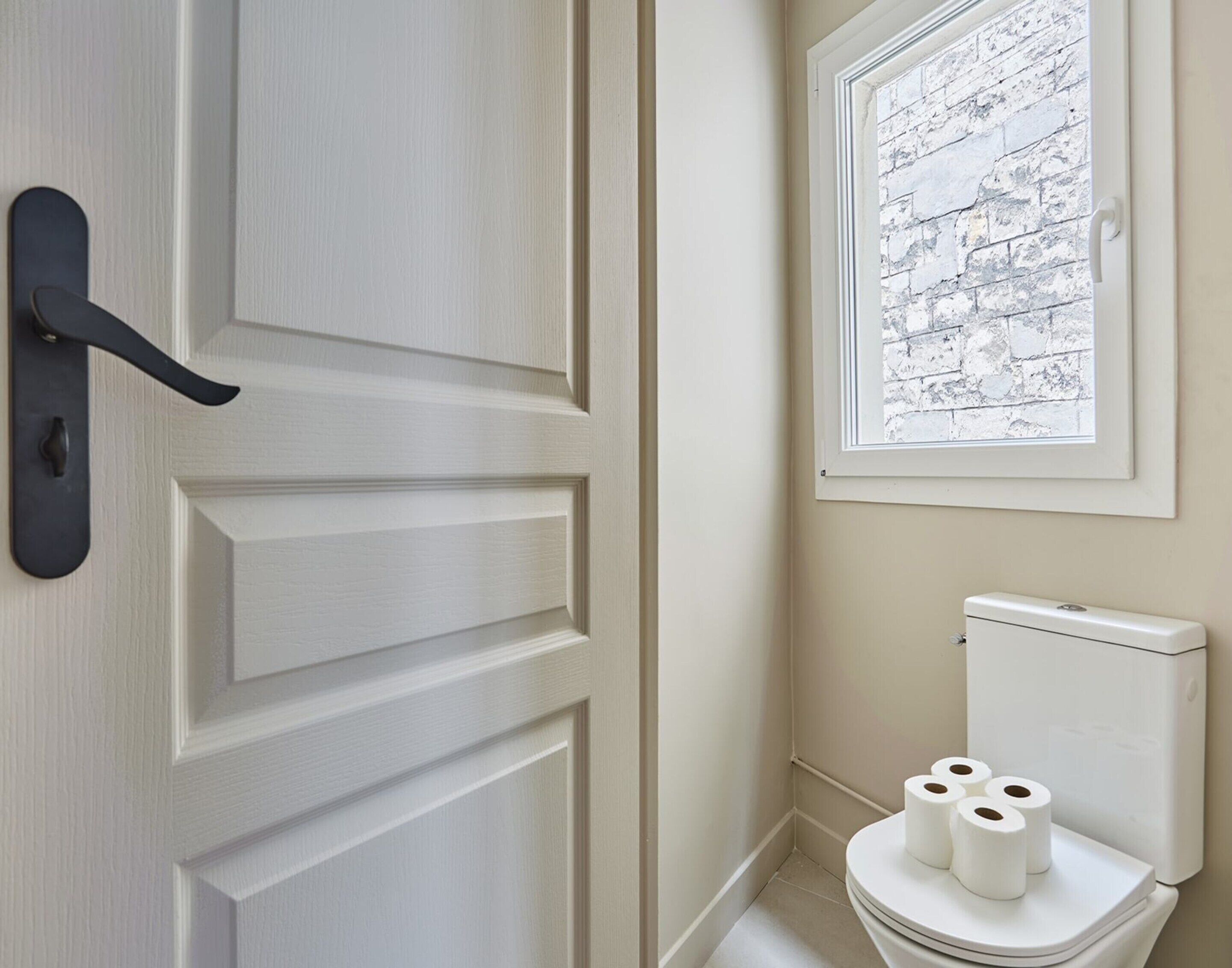 Une salle de bain avec une porte blanche, une fenêtre ornée d’un motif en briques et des toilettes avec du papier toilette.