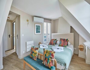 A bedroom with a bed, colorful pillows, a bench, a window, and an air conditioning unit.