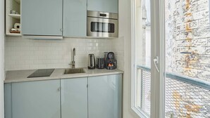 A kitchen with light blue cabinets, a stainless steel oven, a coffee maker, and a window with a view of a brick wall. - Charming Studio Center of Paris Le Marais (Paris)