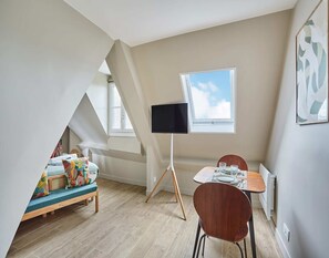 A small, well-lit room with a sloped ceiling, a television on a stand, a dining table with chairs, and a window with a view of the sky.