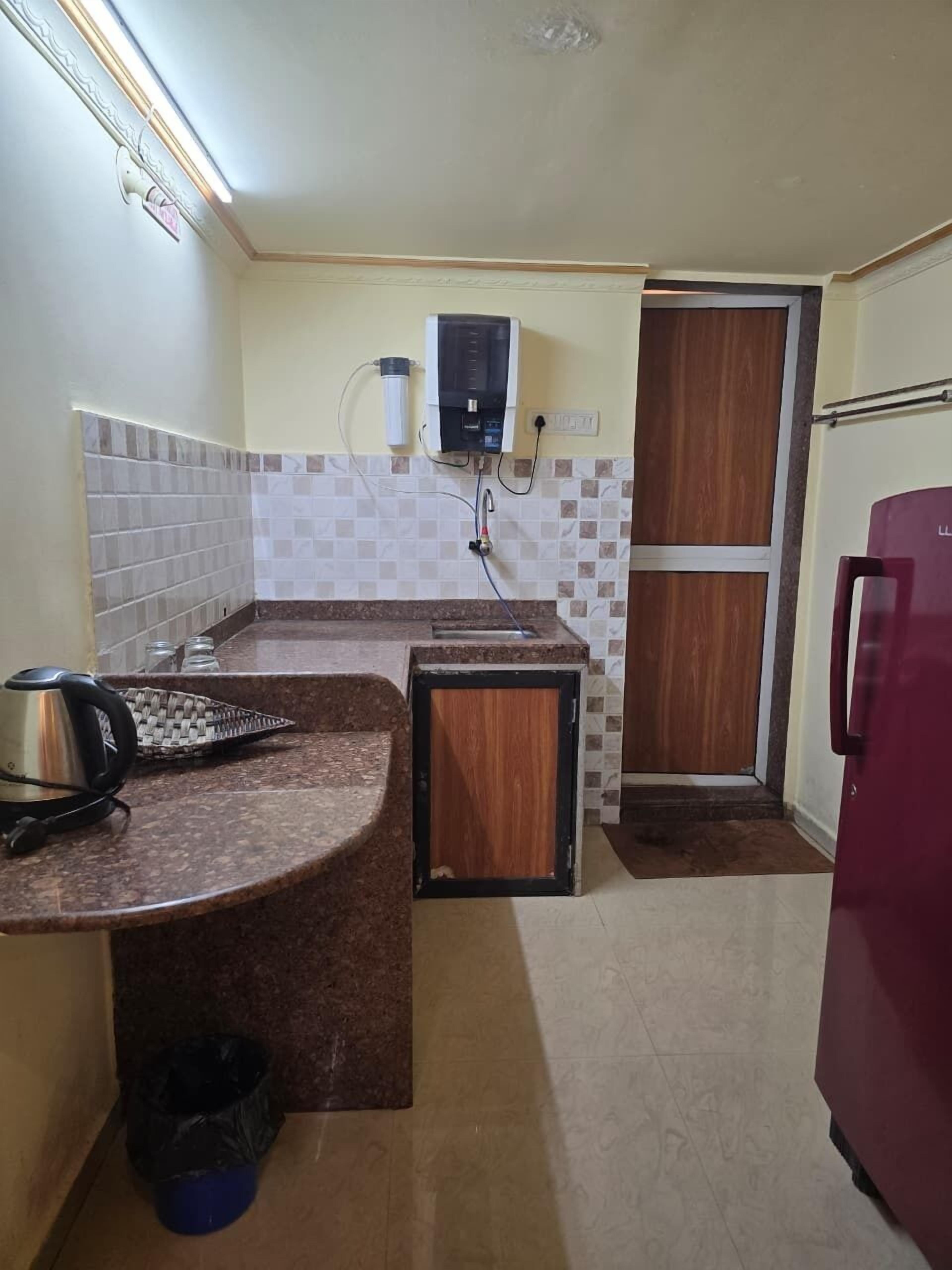 A compact kitchen with a built-in water dispenser, a kettle, and a trash bin.