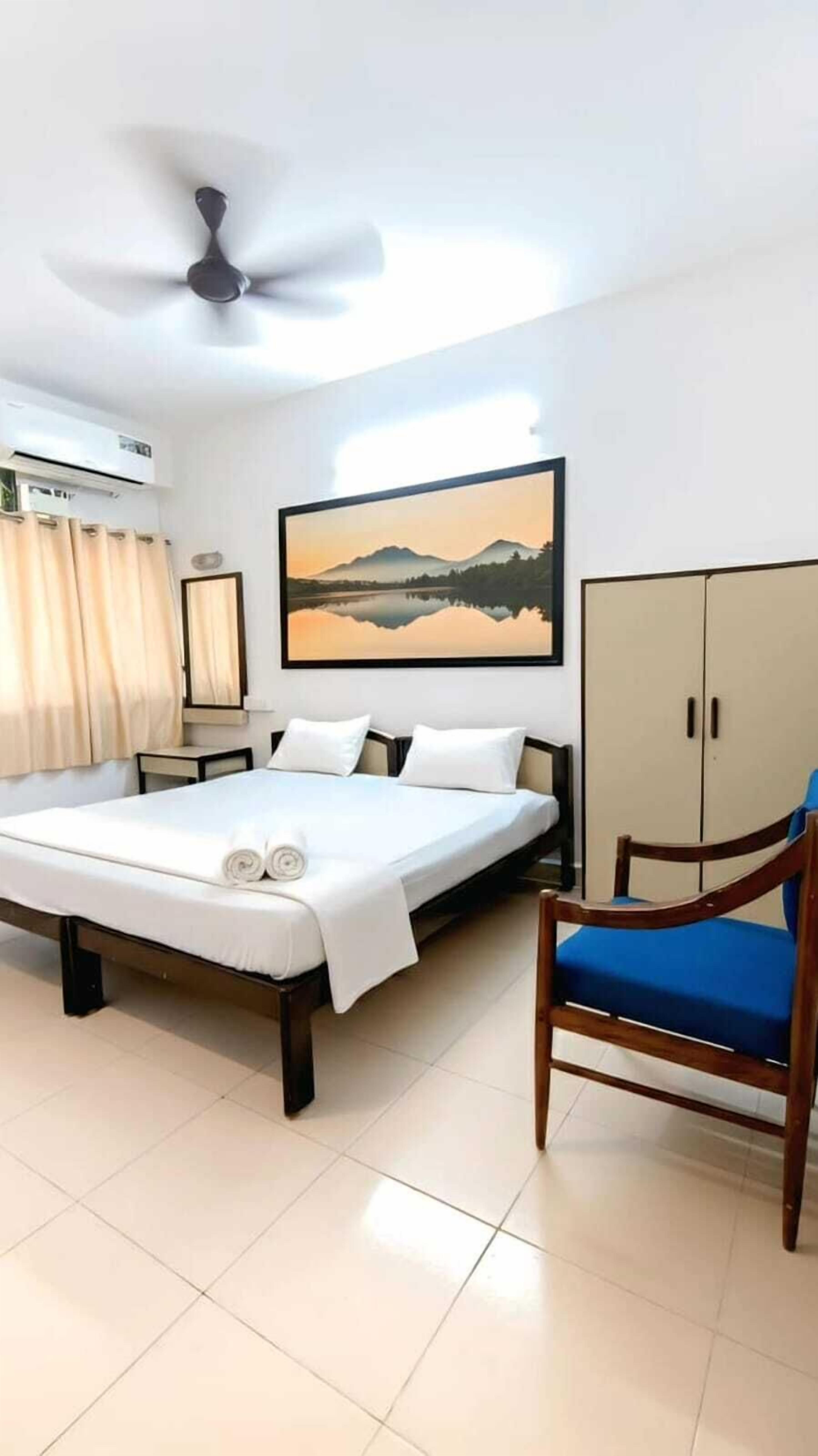 A hotel room with a bed, a chair, a ceiling fan, an air conditioning unit, a painting, and a wardrobe.