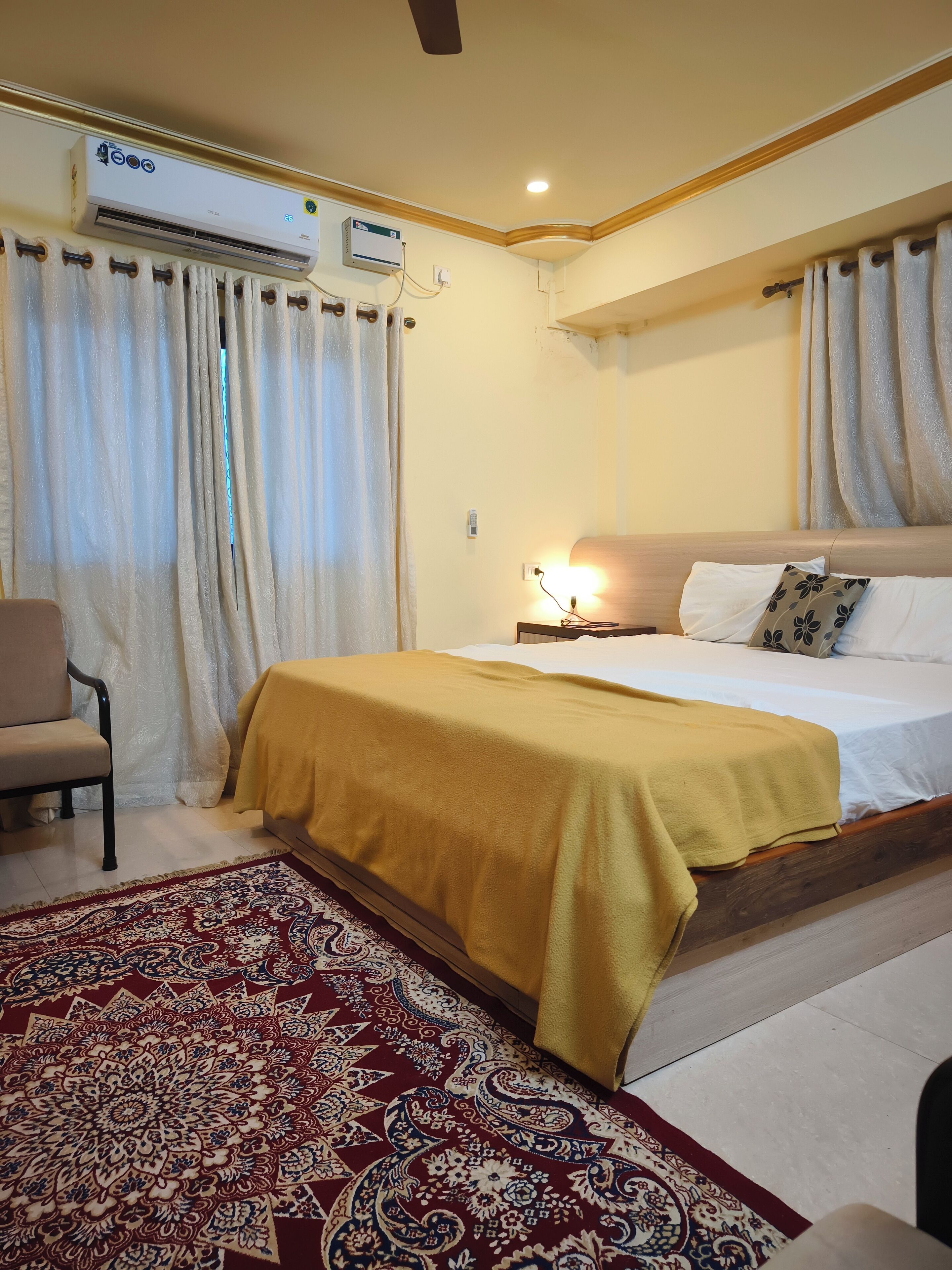 A hotel room with a bed, a chair, a patterned rug, and an air conditioning unit.
