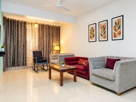 Un salon comprenant un canapé, un fauteuil et une table basse. On y trouve également un ventilateur de plafond, des rideaux et des œuvres d’art encadrées accrochées au mur.