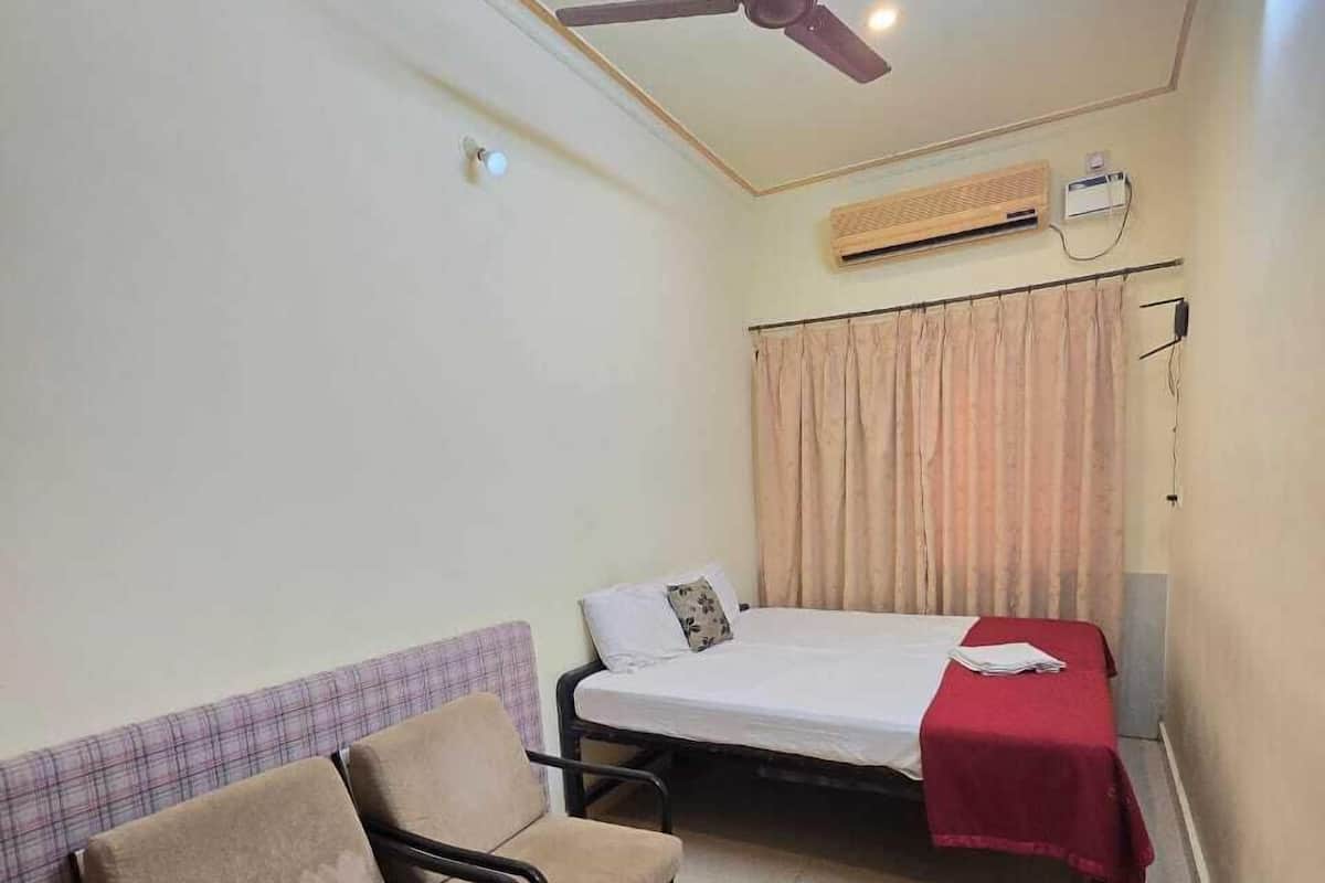 A small, well-lit room with a bed, a chair, a ceiling fan, and an air conditioning unit.