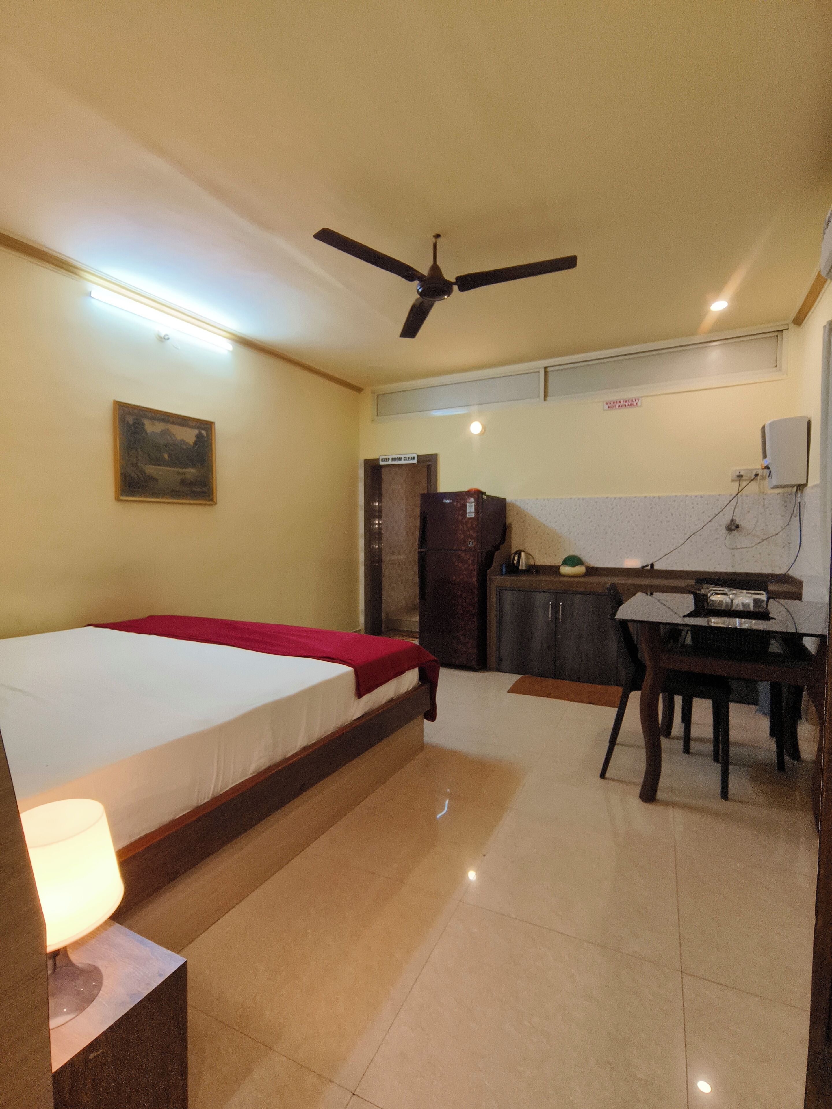 Ein Hotelzimmer mit Bett, Schreibtisch, Stuhl, Deckenventilator, Kühlschrank und Küchenzeile.
