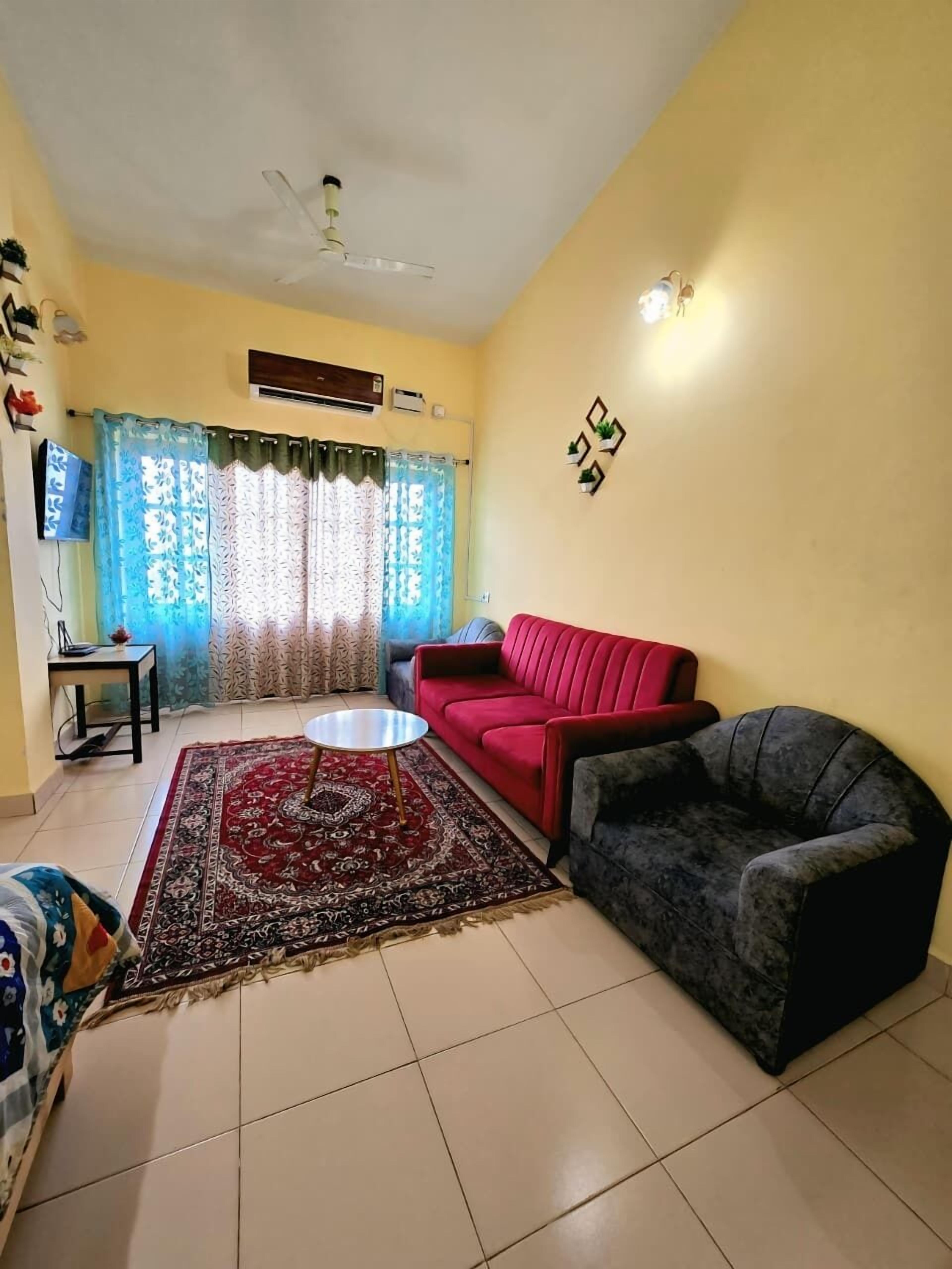 A living room with a red sofa, a grey armchair, a patterned rug, a small round table, and a ceiling fan.