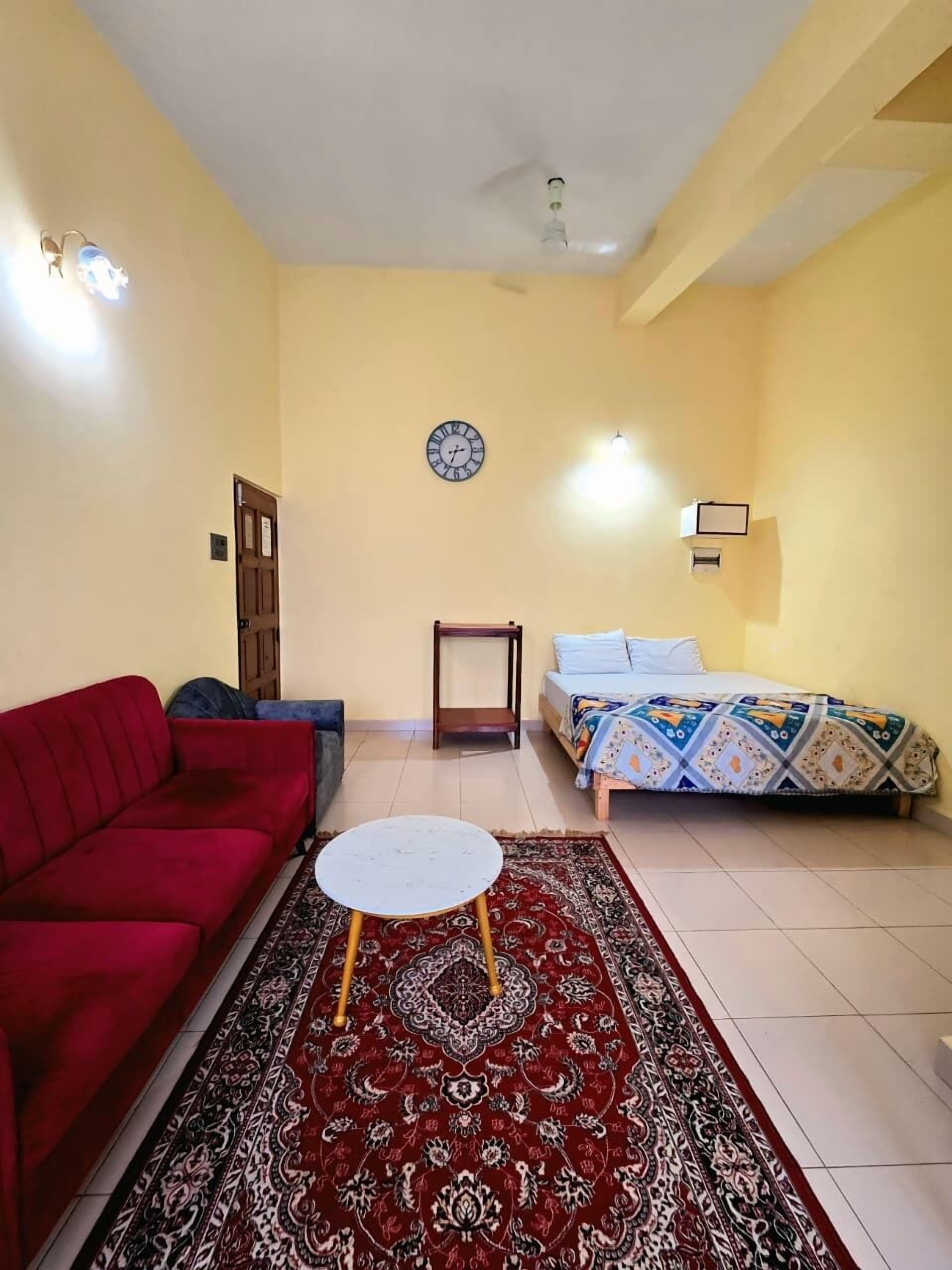 A living room with a red sofa, a small round table, a patterned rug, a bed with a colorful bedspread, a wall clock, and a ceiling fan.