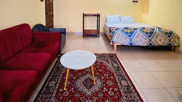 A living room with a red sofa, a small round table, a patterned rug, a bed with a colorful bedspread, a wall clock, and a ceiling fan.