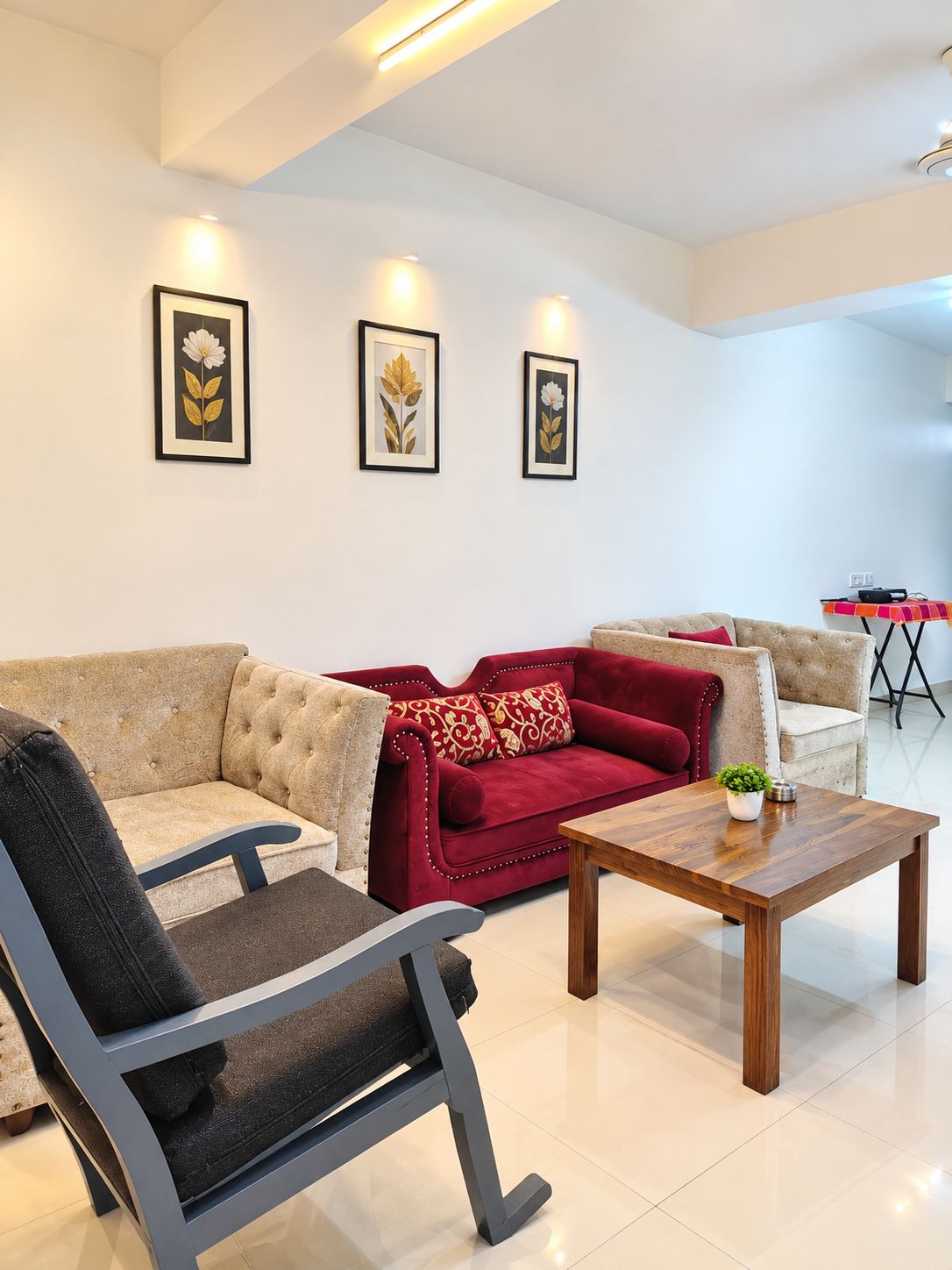 A modern living room with a beige sofa, a red sofa, a wooden coffee table, and a grey armchair. There are three framed artworks on the wall.
