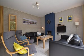 A living room with a sofa, armchairs, a coffee table, a fireplace, a TV, and a clock on the wall. - Fireplace Bungalow in Family Park (Sint Maartensvlotbrug)