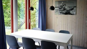 A room with a white table, blue chairs, large windows with red frames, and a wooden wall with a picture. - Exklusives Skandinavisches Ferienhaus (Mirow)