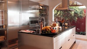 A modern kitchen with stainless steel appliances, a central island with fruits, and a view of greenery through an archway. - RED-HOUSE, Red-House Tenerife Sur (Costa Adeje)