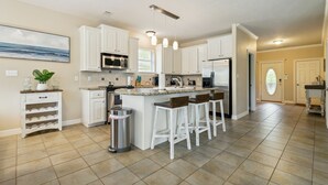 A spacious kitchen with white cabinets, a granite countertop, and a stainless steel refrigerator. There are three bar stools and a trash can.