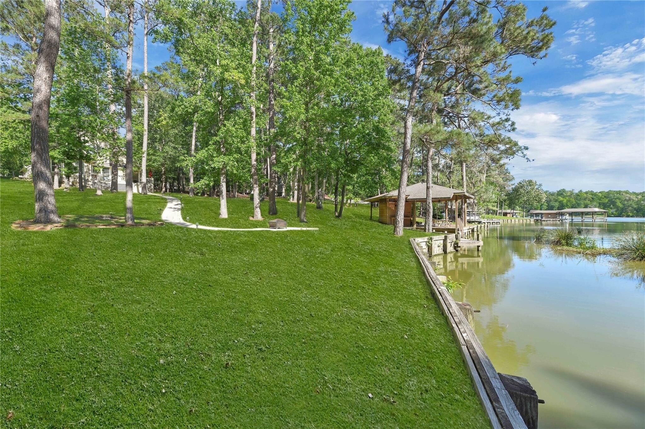 A well-maintained grassy area with a wooden dock extending into a calm lake, surrounded by tall trees and a small wooden structure.