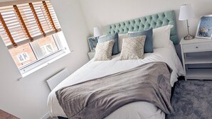 Une chambre à coucher avec une tête de lit capitonnée, un grand lit avec de nombreux oreillers, des tables de chevet avec des lampes et une fenêtre avec des stores.
