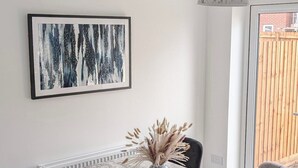 Un coin repas comprenant une table en marbre, quatre chaises, un vase contenant des plantes séchées, un tableau abstrait encadré et un luminaire au plafond.